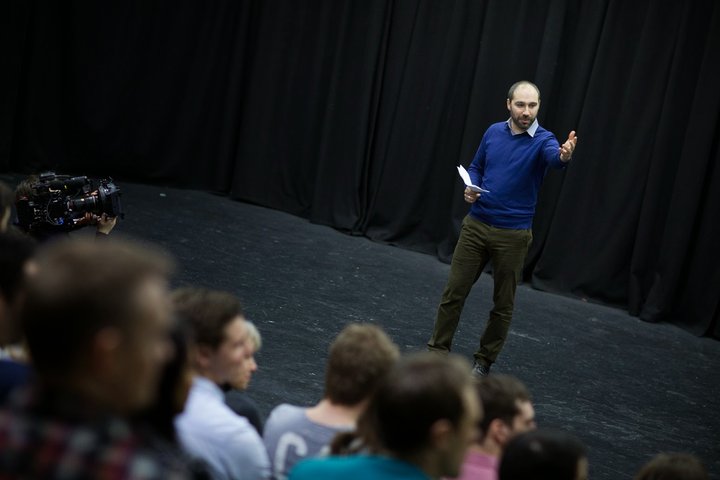 A RADA Business tutor presents to a group of business people in a theatre space. His left arm is outstretched toward the audience.